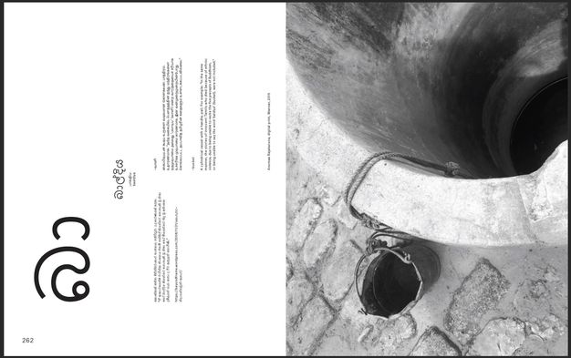 Pages of a book. To our right, a black and white photograph is a view down a well with a bucket tied with a string to our left. The left page has text in English, Sinhala, and Tamil. All are oriented sideways, as if the book opened up instead of out.