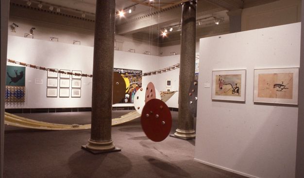 Installation. Three round disks hang from the ceiling between two polished marble columns in a gallery. Works of art and sculpture hang on the floating walls and occupy the space within.