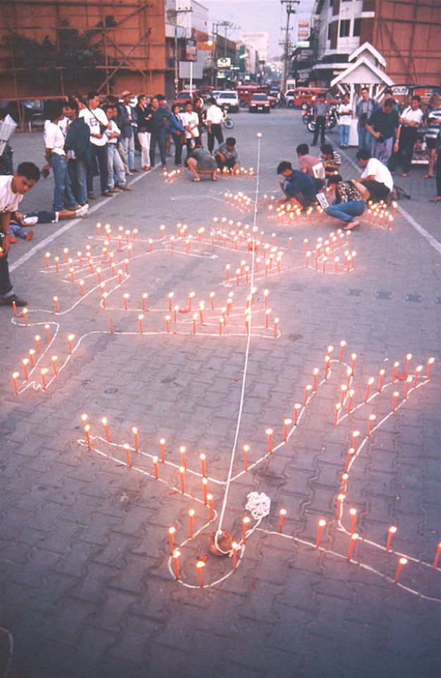 Public art. The outlines of seven people have been outlined in white on a paved sidewalk or street. Orange or red candles are set all around the perimeter of each body. A pile of white string sits inside the chest of the outline closest to us. It unwinds to cut a long line down the centre of the road.