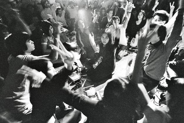 Black and white photograph. About thirty-five people sit or kneel and hold up one hand, first and middle fingers raised to make a V. Cassette players sit on the ground between them.