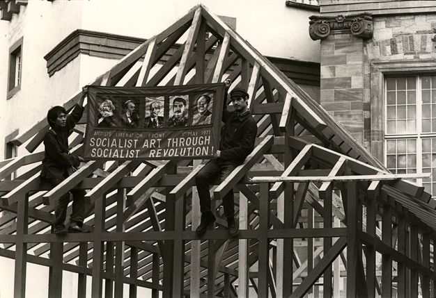 Black and white photograph. Two men hold up a sign reading “Socialist Art through Socialist Revolution!” The men are braced within the rafters of an exposed roof.
