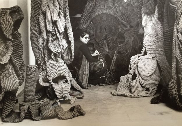 Black and white photograph. A woman sits in a studio draped with woven sculptures.