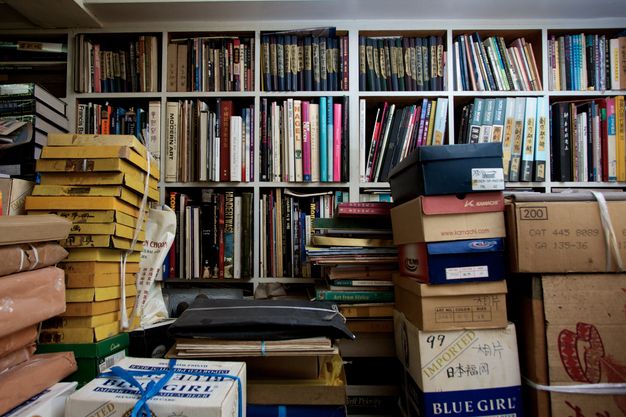 Piles of boxes, files, and flat yellow boxes separate us from the shelves packed with books that fill the wall opposite us.