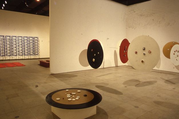 Installation. At least eight round discs, like tabletops, float suspended from a gallery ceiling. A seventh sits on a pedestal and holds round stones or objects made of another material. The hanging discs are painted brown, red, or black. A sculptural piece hanging on the wall seen through a doorway to our left is of ladder or trellis-like structures creating a dense geometric pattern.