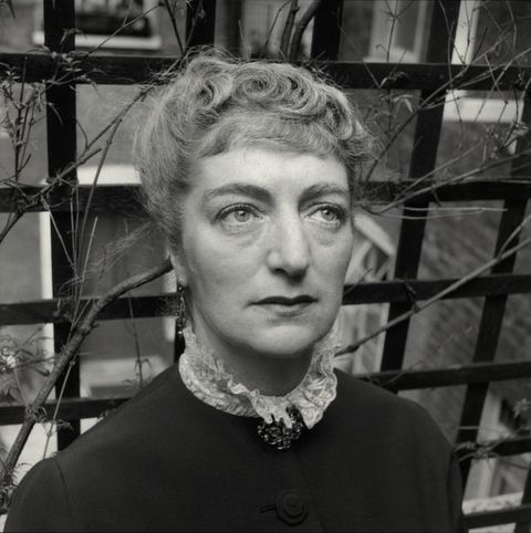 Black and white photograph. Shown from the shoulders up, a pale-skinned woman looks up and to our left. She has a high lace collar over a high-necked jacket, pinned at the throat with a brooch. Her curly hair is pulled up. She stands in front of a trellis with twiggy vines.