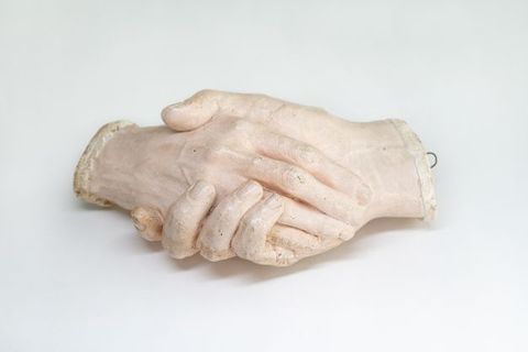 Sculpture. Plaster cast of two right hands holding each other. The hands cut off at the wrists. In this view, a hand rests along the palm of an encasing hand.
