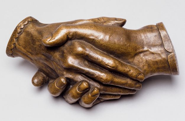 Sculpture. Bronze cast of two right hands holding each other. The hands cut off at the wrists, and one is edged in a straight cuff and the other in scalloped cuff. In this view, one rests along the palm of the other. Veins on the back of one hand and on the underside of the wrist of the other are visible. The patina is honey brown.