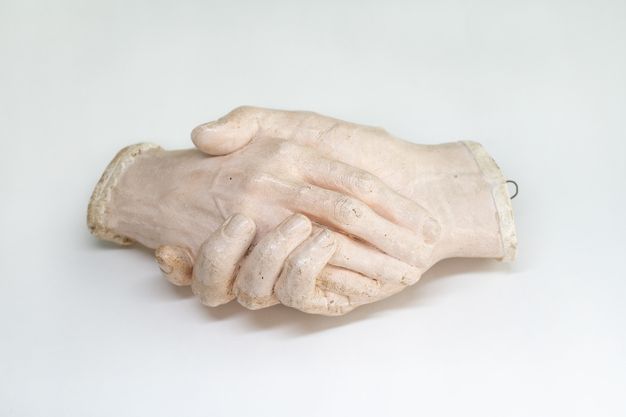Sculpture. Plaster cast of two right hands holding each other. The hands cut off at the wrists. In this view, a hand rests along the palm of an encasing hand.