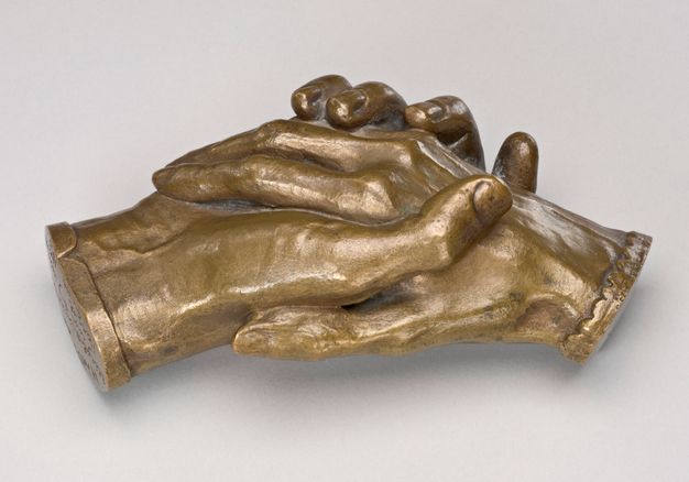 Sculpture. Bronze cast of two right hands holding each other. The hands cut off at the wrists, and one is edged in a straight cuff and the other in scalloped cuff. In this view, the thumbs tuck in against each other. Veins on the back of one hand and on the underside of the wrist of the other are visible. The patina is an almost flat gold colour.