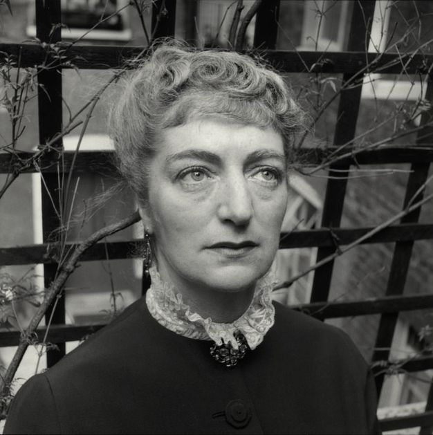 Black and white photograph. Shown from the shoulders up, a pale-skinned woman looks up and to our left. She has a high lace collar over a high-necked jacket, pinned at the throat with a brooch. Her curly hair is pulled up. She stands in front of a trellis with twiggy vines.