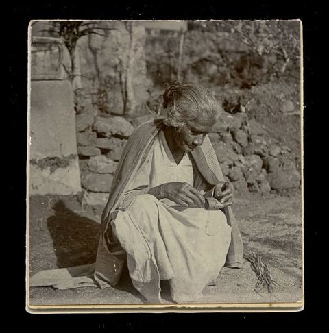 Black and white photograph. Eight people stand on either side of long threads that stretch across at least three thatched houses at waist height. One person bends over the threads. All wear cloth wrapped around their hips or draped over their shoulders. A pencil inscription below the image reads, “Plate I, Weavers”.