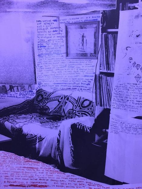 Installation. A black and white photograph tinted delphinium blue is of a room with a blanket-covered couch. Red or blue text has been written on the walls and floor.