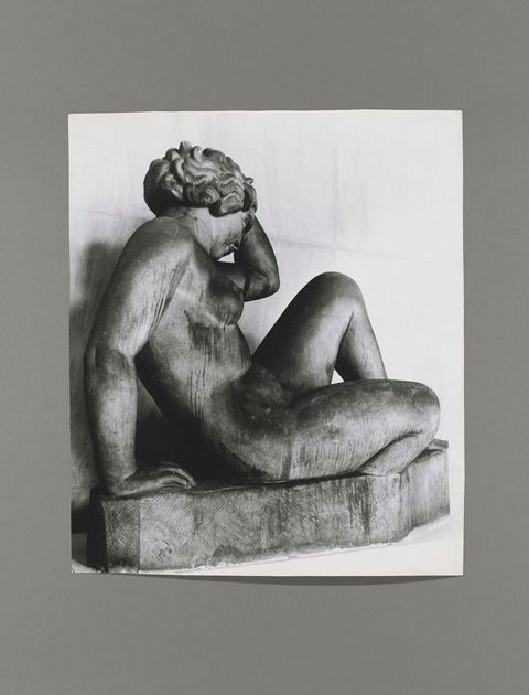 Black and white photograph. A sculpture of a nude woman, her features and hair simplified and stylized, leaning back on a tall base. Her body is angled to our right and slightly away from us. One hand is lifted to wavy hair, the other supports her body. The surface is dark and streaked.