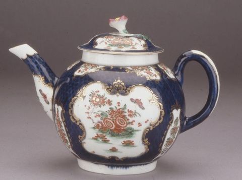 Teapot. A round-bodied ceramic teapot has an upwards lilting handle opposite the spout, which is to our left in this view. The body and lid are painted with brick-red flowers and butterflies in white cartouches, surrounded by gold edging against a navy-blue background. A pink-tipped flower sprouts from the lid.