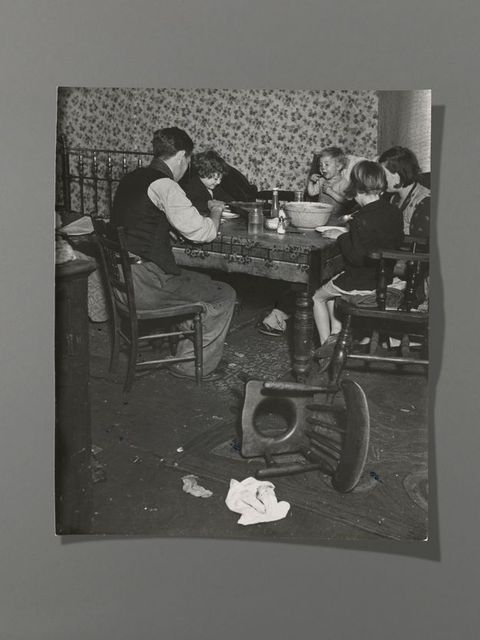 Black and white photograph. A man, woman, two children, and a baby gather around a dining table. Most have their backs to us, but one child and the baby face us, smiling. An overturned commode chair is between us and the group.