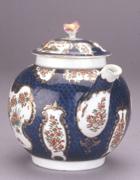 Teapot. The spout of a round-bodied ceramic teapot comes towards us, angled to our right, in this view. The body and lid are painted with brick-red flowers and butterflies in white cartouches, surrounded by gold edging against a navy-blue background. A pink-tipped flower sprouts from the lid.