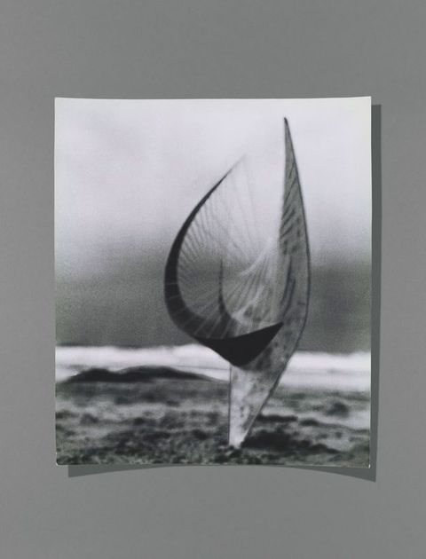 Black and white photograph. A sculpture rising from a sandy beach is blurry against the even blurrier breaking surf beyond. The sculpture seems to rise from a point in the sand to flare out into a fractured, bowl-like U shape, with strings connecting different parts.
