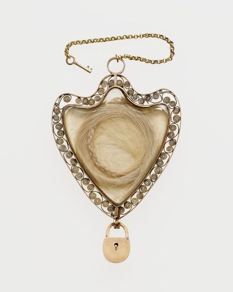 Jewellery. A shield-shaped pendant contains a braided locket of pale hair. The edge is surrounded by scrolling filigree. A miniature padlock hangs from the bottom and a tiny skeleton key is attached by a chain to the top.