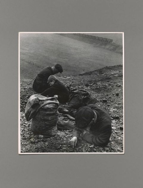Black and white photograph. Two men and a boy sit or lean against a steep incline, picking through dirt and pebbles. A sack sits next to the man closest to us. A flat field beyond the incline leads back to a train in the distance.