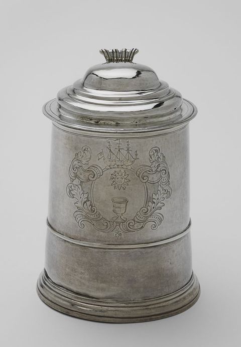 Tankard. A silver, lidded vessel is incised with imagery surrounded by strapwork and foliation, like a heraldic crest. The disc in the centre shows a goblet under a sun. A three-masted ship is above the disc.