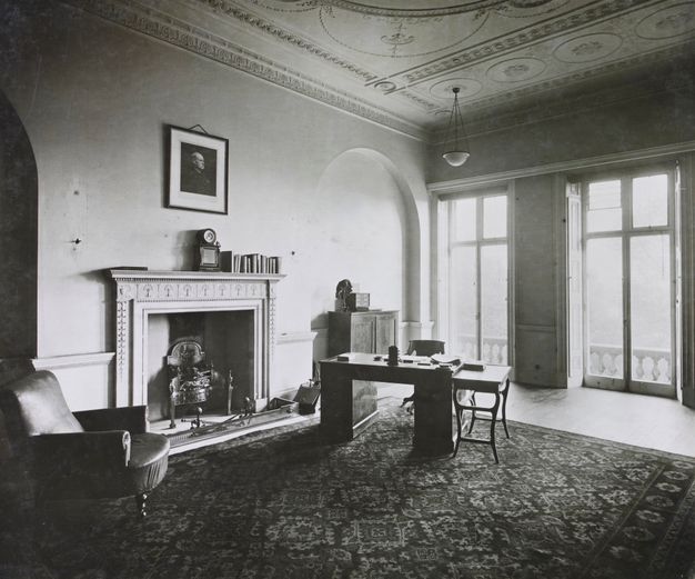 Black and white photograph. A high-ceiling room is furnished sparsely with a patterned rug, a small desk and table, a cabinet, and an armchair. About a dozen books are lined up on the mantle below a portrait hanging above. The mantle and ceiling are decorated with plasterwork, and windows across from us span nearly the height of the walls.