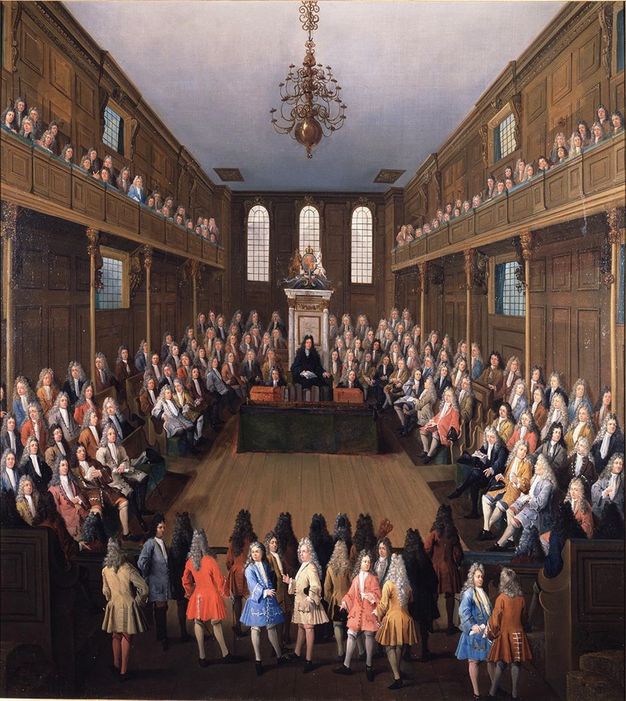 Painting. A hall-like, wood-panelled room is filled with dozens of pale-skinned men wearing white, grey, or black wigs and suits in coral red, ultramarine blue, rose pink, deep yellow, grey, brown, or cream white. They sit in rows along the walls and on a balcony level, and others stand.