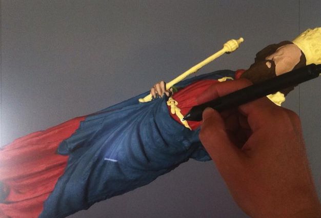A hand holding a digital pen adds yellow to a computer model of a sculpture. The statue is of a pale-skinned man with a brown beard and hair, red and blue robes, and a yellow crown, sceptre, orb, and belt.