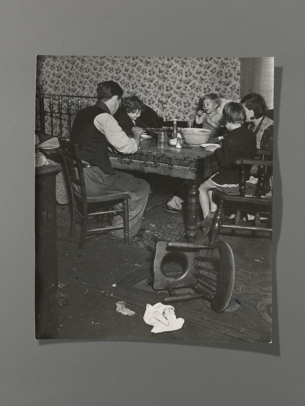 Black and white photograph. A man, woman, two children, and a baby gather around a dining table. Most have their backs to us, but one child and the baby face us, smiling. An overturned commode chair is between us and the group.