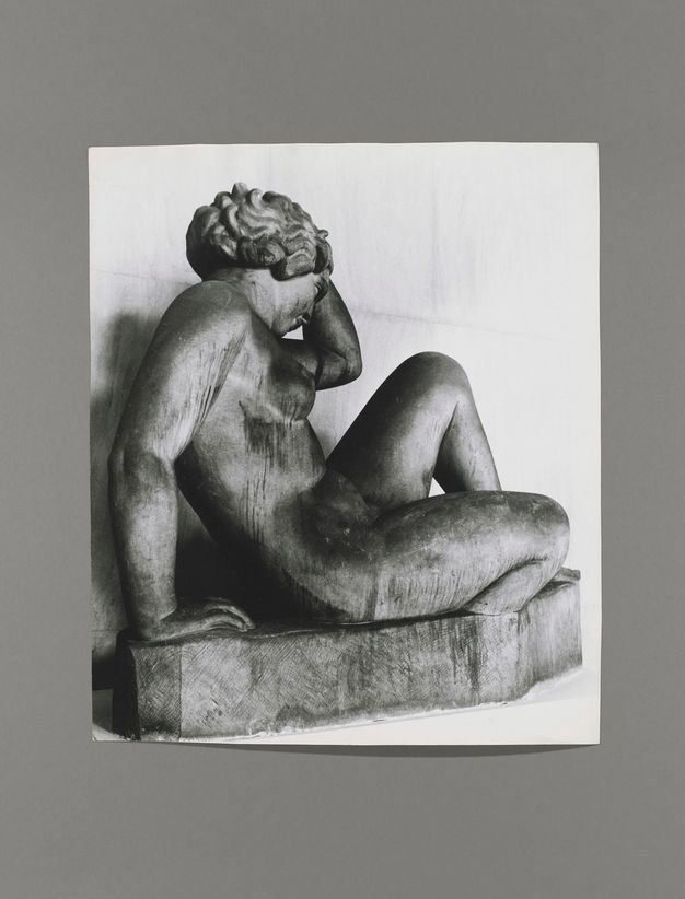 Black and white photograph. A sculpture of a nude woman, her features and hair simplified and stylized, leaning back on a tall base. Her body is angled to our right and slightly away from us. One hand is lifted to wavy hair, the other supports her body. The surface is dark and streaked.