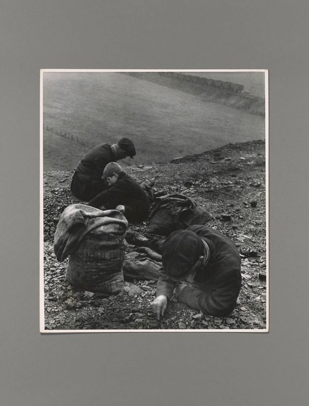 Black and white photograph. Two men and a boy sit or lean against a steep incline, picking through dirt and pebbles. A sack sits next to the man closest to us. A flat field beyond the incline leads back to a train in the distance.
