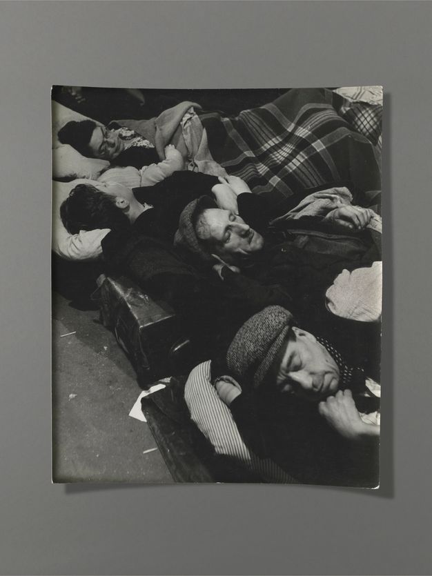 Black and white photograph. Men, women, and a baby sleeping fully clothed, shoulder to shoulder, fill this composition.
