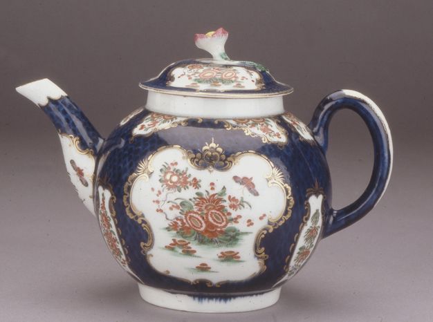 Teapot. A round-bodied ceramic teapot has an upwards lilting handle opposite the spout, which is to our left in this view. The body and lid are painted with brick-red flowers and butterflies in white cartouches, surrounded by gold edging against a navy-blue background. A pink-tipped flower sprouts from the lid.