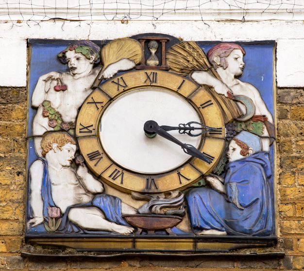 Architectural detail. A round clock face on the exterior of a building is set into a larger square, the corners of which are filled with personifications of the seasons. Children with white-coloured skin hold berries or sheaves of wheat, or sit by a fire or next to a flower. Most of the glazes are vivid blue or golden yellow, with a few touches of red and green.