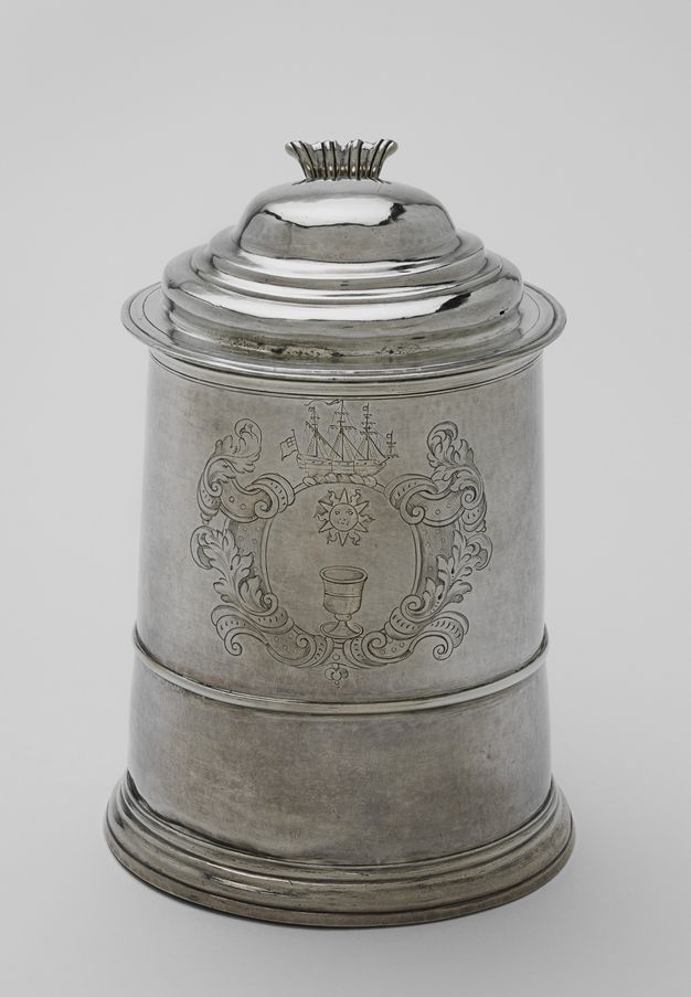 Tankard. A silver, lidded vessel is incised with imagery surrounded by strapwork and foliation, like a heraldic crest. The disc in the centre shows a goblet under a sun. A three-masted ship is above the disc.