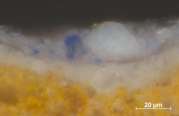 Microscopic cross section of paint. From the bottom up are layers of golden yellow, white, and finally, a layer of lapis blue, white, and traces of dark pink. Text in the lower right reads, “20 µm”.