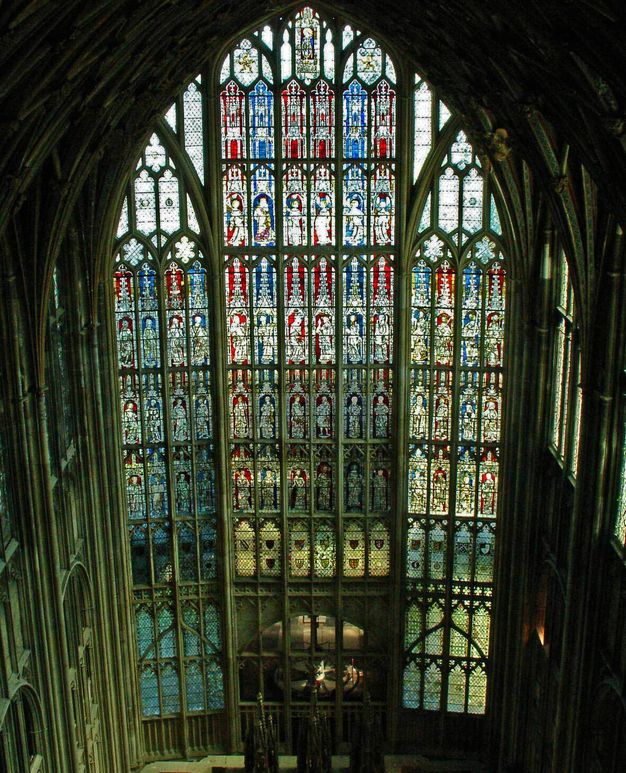 Architecture. We look down onto a stained glass window that fills an entire wall. Pointed arches nested within arches create an intricate, lacy design. The windows are filled with blue, red, and clear glass.