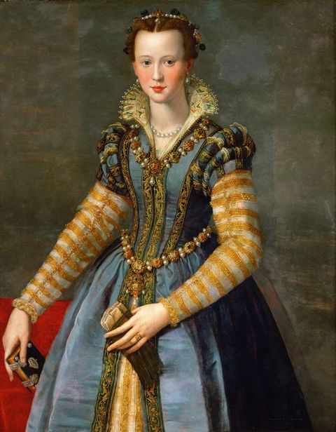 Painting. Shown from the knees up, a pale-skinned woman’s cheeks flush pink and her red bow-shaped lips slightly smile. A pearl diadem tucks into her auburn hair, which is pulled back. Her gown is laden with gems and pearls in gold fittings and with gold-edged and patterned fabric. She holds a postcard-sized book in one hand and a pair of gloves in the other.