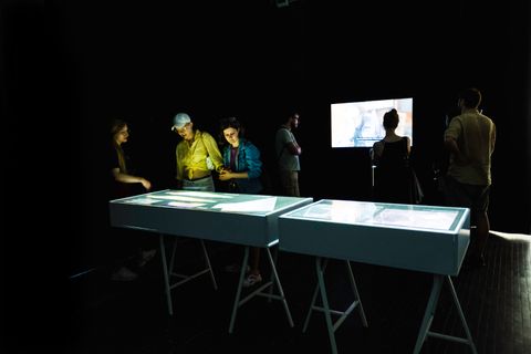 Installation. In a darkened room six people look at images in display cases or watch a video on the wall opposite us.