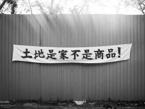 Mixed media. A black and white photograph sun streaking over the top edge of a solid fence holding a wide white banner. Chinese characters have been written across the banner.