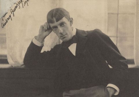 Black and white photograph. Shown from the waist up, a pale-skinned man with a long, narrow face sits with one elbow propped on a window ledge behind him. His hair is parted down the middle and cut into straight bangs across his high forehead.