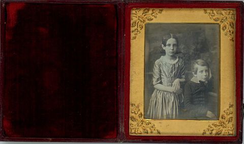 Daguerreotype in an open case. A young girl stands and a boy sits, and they look out at us. They both have pale skin and prop their left elbows, to our right, on ledges behind them. They are framed in the rectangular opening of a gold mat, which has leafy designs at the corners. The case flips up, and the cover is lined with deep red fabric.
