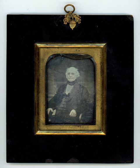 Daguerreotype. A pale-skinned, balding man with a deeply lined face sits looking off to our left. The rectangular gold mat is arched on the upper corners. A wide black frame surrounds it, and a gold acorn is at the top centre.