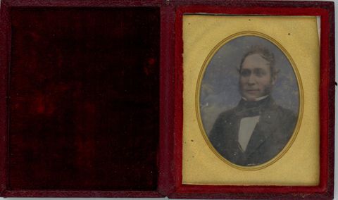 Daguerreotype in an open case. A pale-skinned man looks off to our left. His cheeks and lips have been tinted pink, and the background tinted blue. He is framed in the oval opening of a gold mat. The lid of red case is flipped open to our left and is lined with dark red cloth.