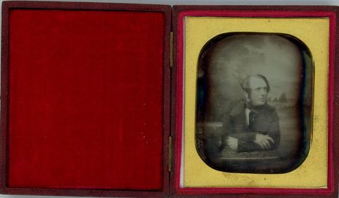 Daguerreotype. A pale-skinned man with mutton chops leans forwards onto his crossed arms as he looks off to our right. His dark hair is swept forwards over his ears, and his lips are parted. The background shows a distant landscape.