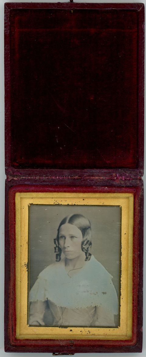 Daguerreotype in an open case. A light-skinned woman with curls framing her face looks off to our left. Pink has been added to her cheeks and dress beneath a white shawl. She is shown in a rectangular gold mat. The case flips up and the cover is lined with dark red fabric.
