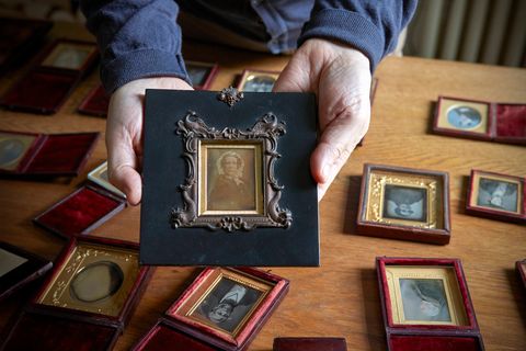 Colour photograph. Two hands hold a framed daguerreotype portrait of a woman wearing a lacy bonnet. The frame around the gold mat is raised in a design of birds and stylized fish. More daguerreotypes are laid out on a tabletop behind the hands.