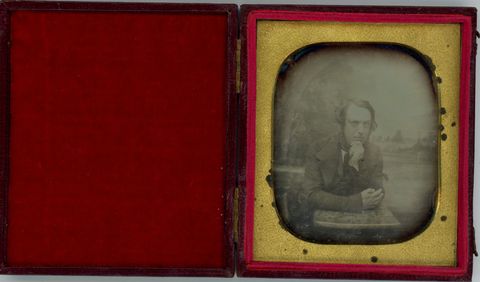 Daguerreotype. A pale-skinned man with mutton chops leans forwards onto his elbows, his chin resting in one hand. He looks at us from in front of a distant landscape background. The gold mat is rounded at the corners.