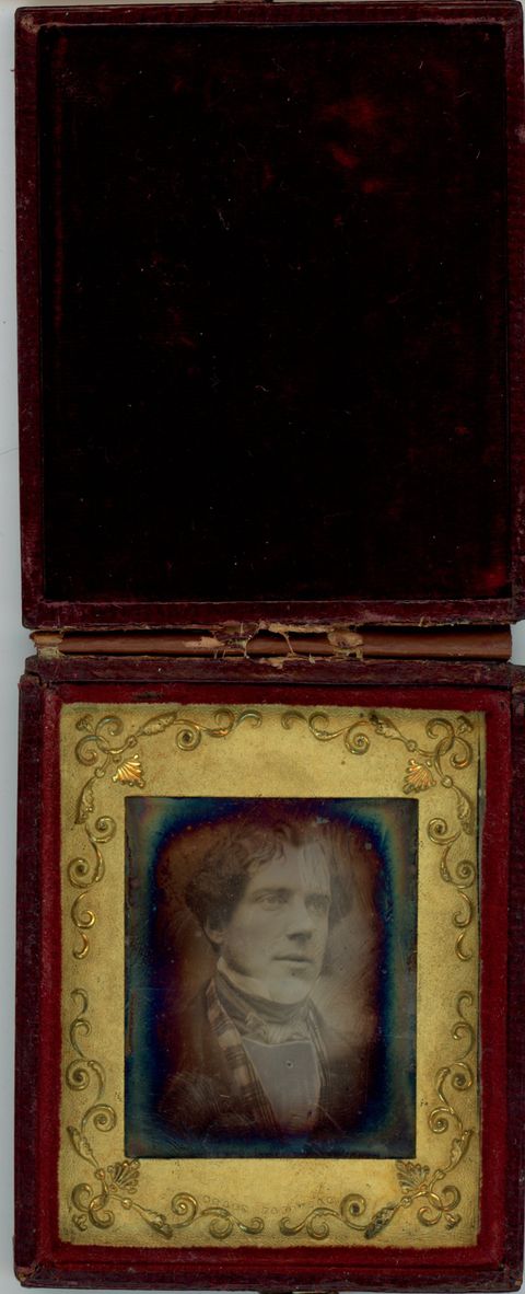 Daguerreotype in an open case. A pale-skinned man with dark hair and short mutton chops looks off to our right. The edges of the photograph are rainbow coloured, like oil on water. He is framed in the rectangular opening of a gold mat, which is decorated with scrolling vines. The lid of the case is flipped up, and the underside is lined with dark red fabric.