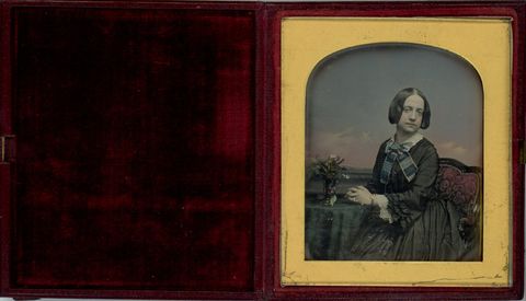 Daguerreotype. A pale-skinned woman wearing a black dress looks at us. Her hands are clasped and rest on a table holding a spray of flowers in a vase. Hand-colouring has been added to the props, background, clothing, and her skin.