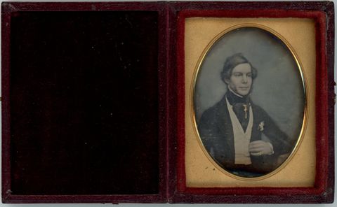 Daguerreotype in an open case. A pale-skinned man with mutton chops sits looking off to our right, a slight smile playing on his wide mouth. The background is tinted blue and his cheeks pink. A jewel on the puffy cravat at his throat may be coloured yellow.
